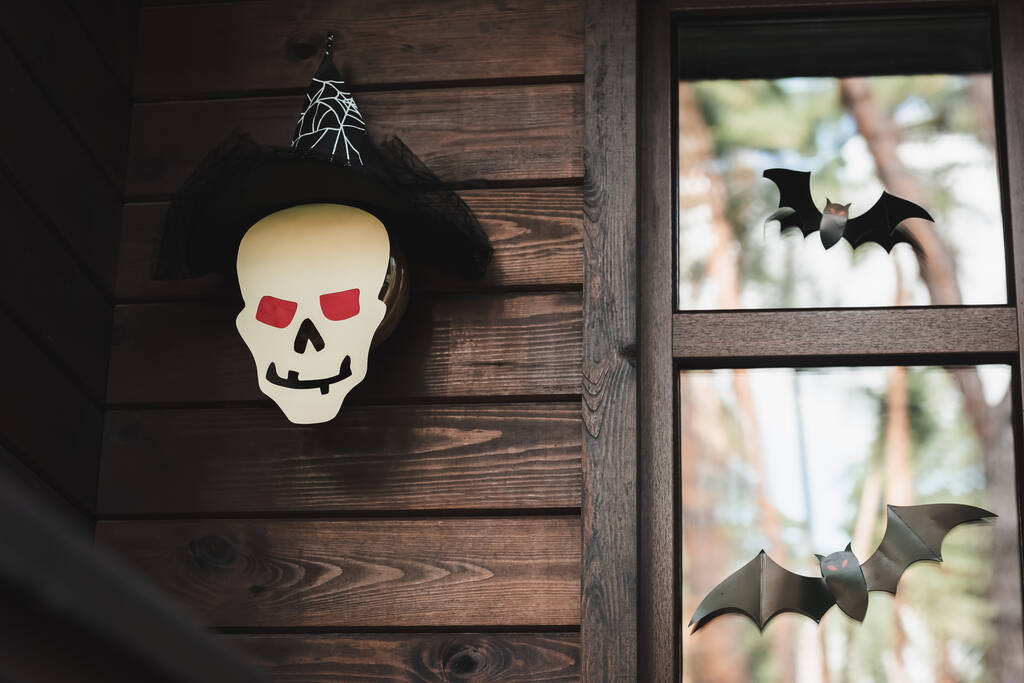 бумага разрезает череп в колпаке ведьмы возле черных летучих мышей на окне деревянного коттеджа - Фото, изображение