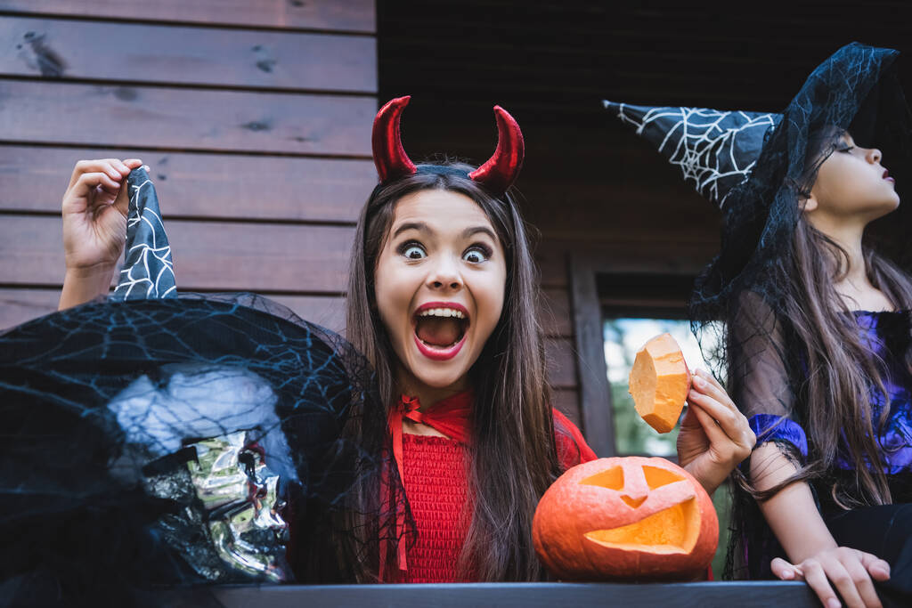 изумленная девушка в костюме дьявола рядом с сестрой в колпаке ведьмы и вырезанной тыкве на крыльце - Фото, изображение