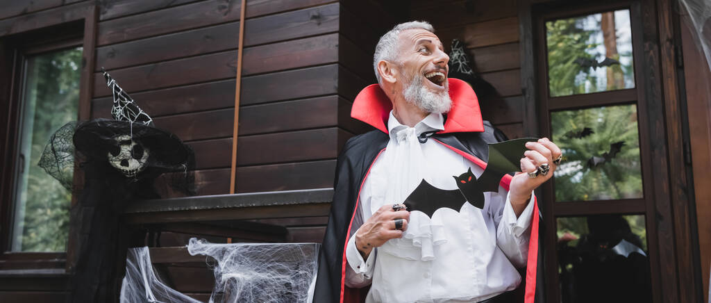 lachen middelbare leeftijd man in halloween vampier kostuum met papier gesneden vleermuis op veranda van houten huis, banner - Foto, afbeelding