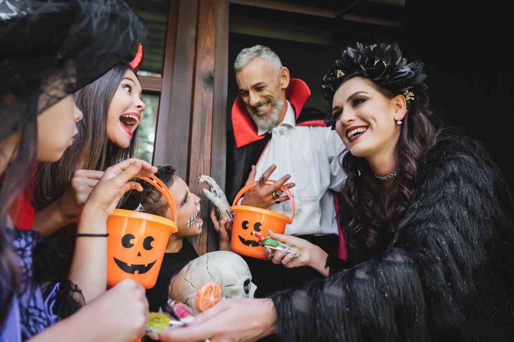 gelukkig paar in halloween kostuums geven snoep aan opgewonden kinderen met emmers - Foto, afbeelding
