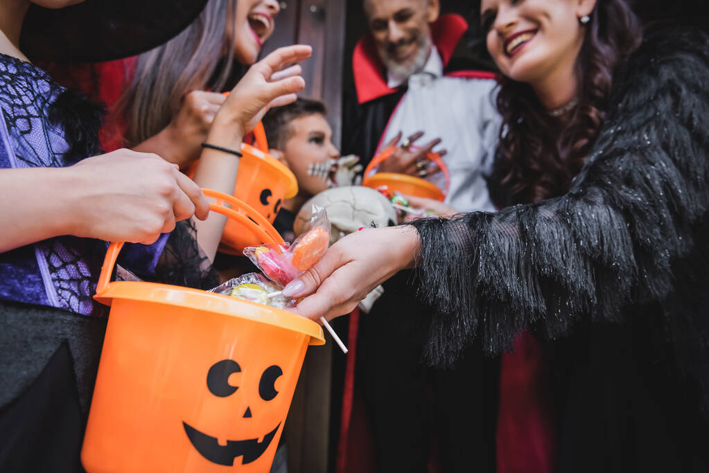 Улыбающаяся женщина кладет конфеты в ведро на Хэллоуин рядом с размытыми детьми - Фото, изображение