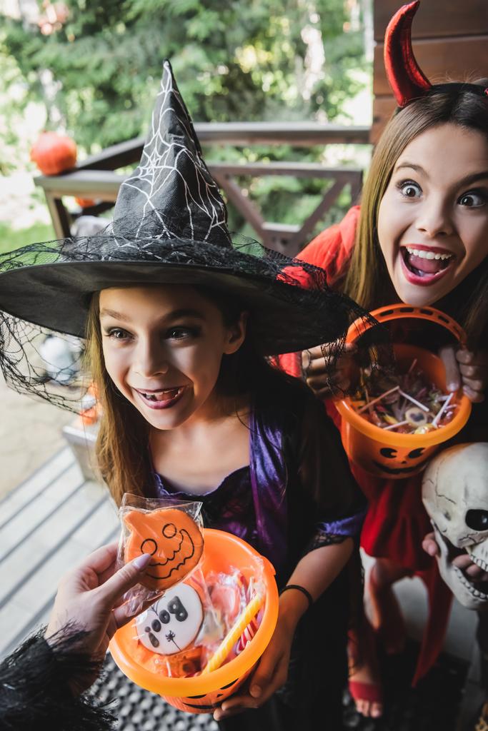 irmãs espantadas em trajes de bruxa e diabo perto da mãe com biscoito de Halloween - Foto, Imagem