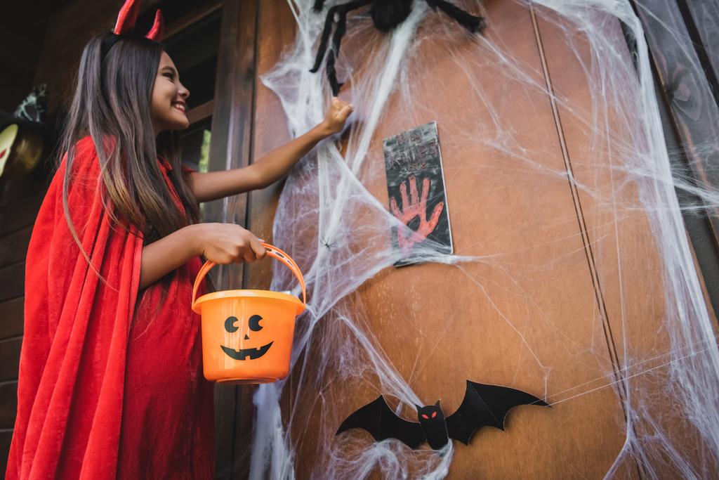 Şeytan kostümü giymiş neşeli bir kızın cadılar bayramı süsüyle kapıyı çalması. - Fotoğraf, Görsel