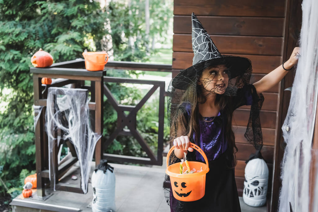 bambino in costume strega Halloween che tiene secchio con dolci mentre bussa alla porta sul portico decorato - Foto, immagini