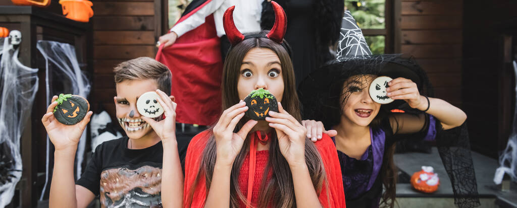 dzieci w upiornych kostiumach trzymających ciasteczka halloween w pobliżu twarzy, baner - Zdjęcie, obraz