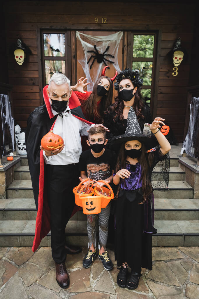 család fekete orvosi maszkok és ijesztő jelmezek tartó vödrök édességek és faragott tök közelében ház - Fotó, kép