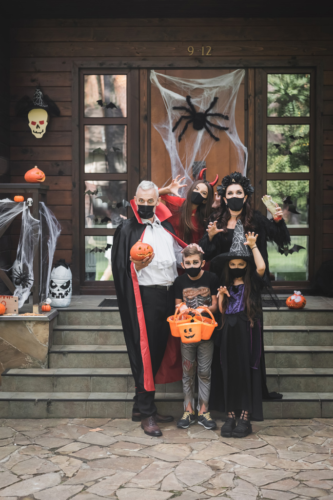 przerażająca rodzina w halloween kostiumy i czarne maski medyczne pokazując straszne gesty w pobliżu ganku z dekoracji - Zdjęcie, obraz