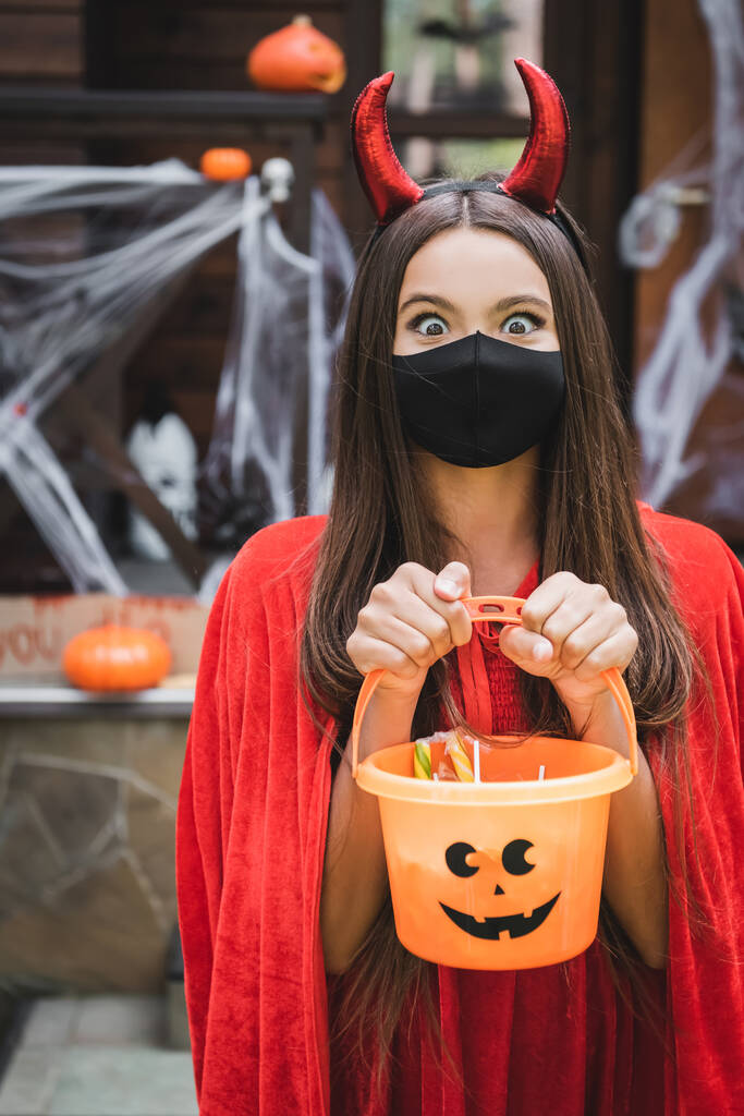 aufgeregtes Mädchen in Teufelskostüm und schwarzer medizinischer Maske, das einen Eimer mit Süßigkeiten in die Kamera hält - Foto, Bild