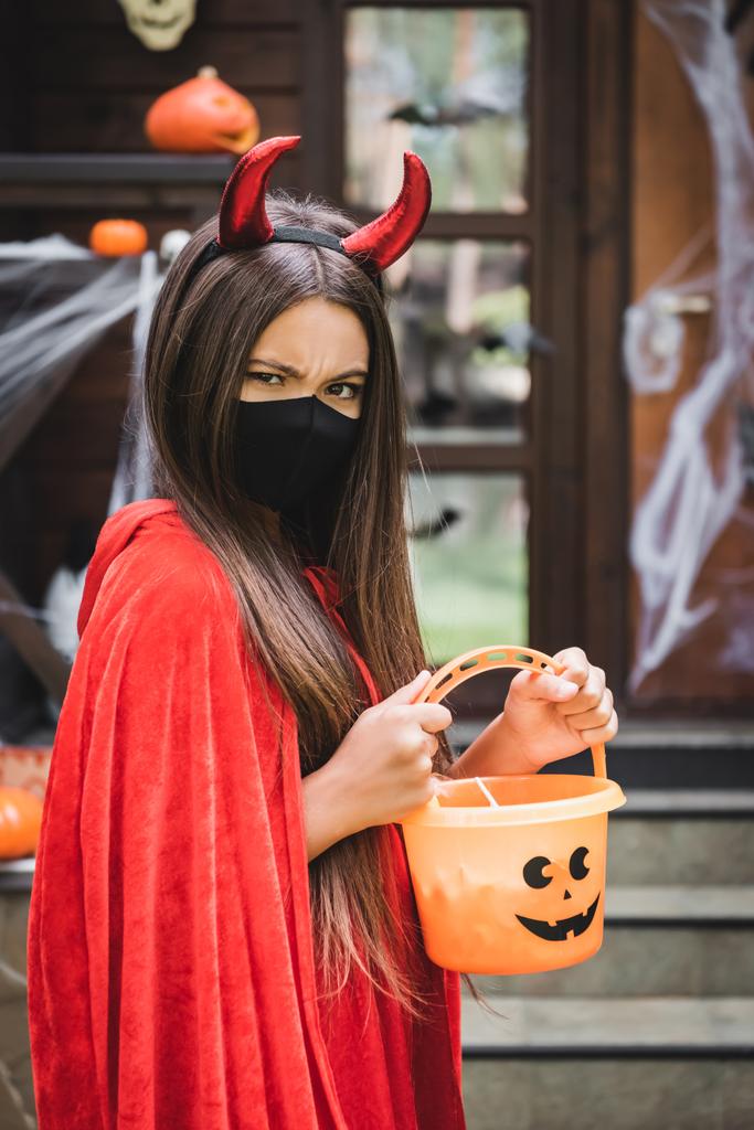 menina zangada em traje de Halloween diabo e máscara médica preto segurando balde de doces perto de casa turva - Foto, Imagem