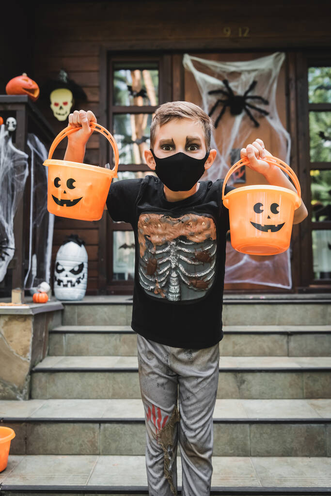 niño en traje de esqueleto y máscara médica negro sosteniendo cubos de Halloween cerca de cabaña borrosa - Foto, Imagen