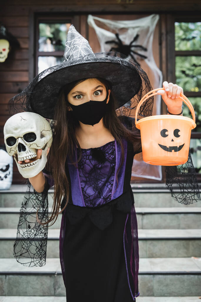 girl in witch halloween costume and black medical mask holding skull and bucket near blurred house - Photo, Image