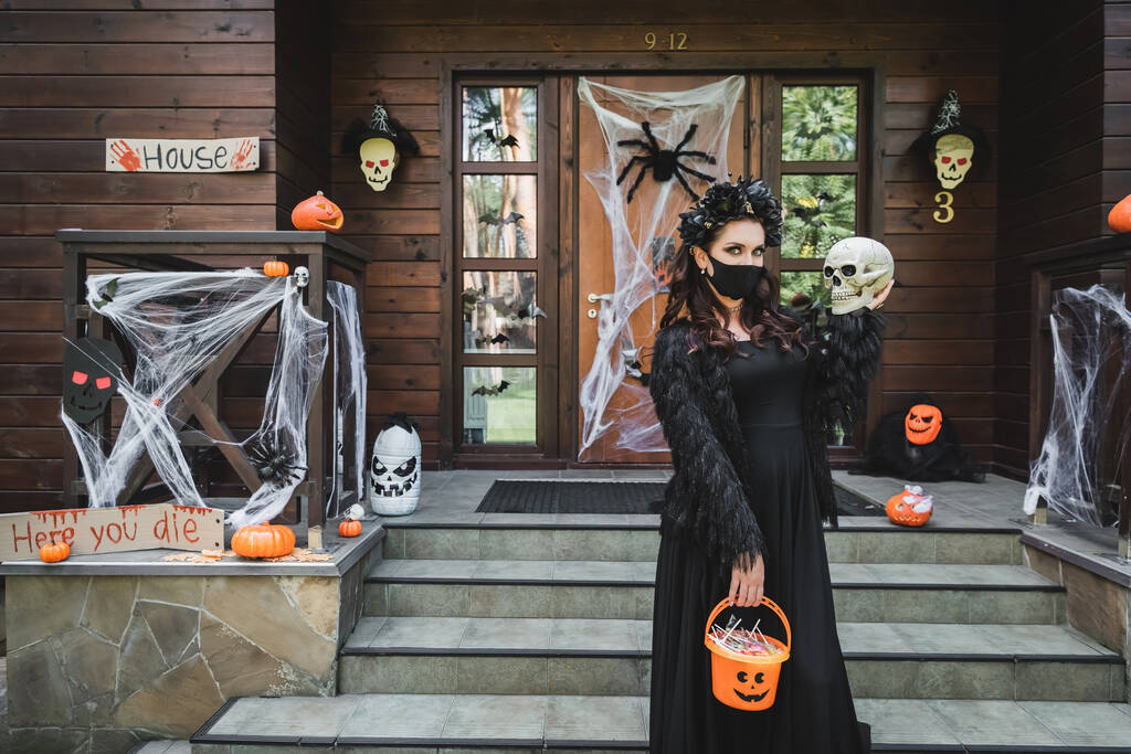 Frau in Halloween-Kostüm und schwarzer medizinischer Maske steht mit Totenkopf und Eimer voller Süßigkeiten in der Nähe der geschmückten Veranda - Foto, Bild