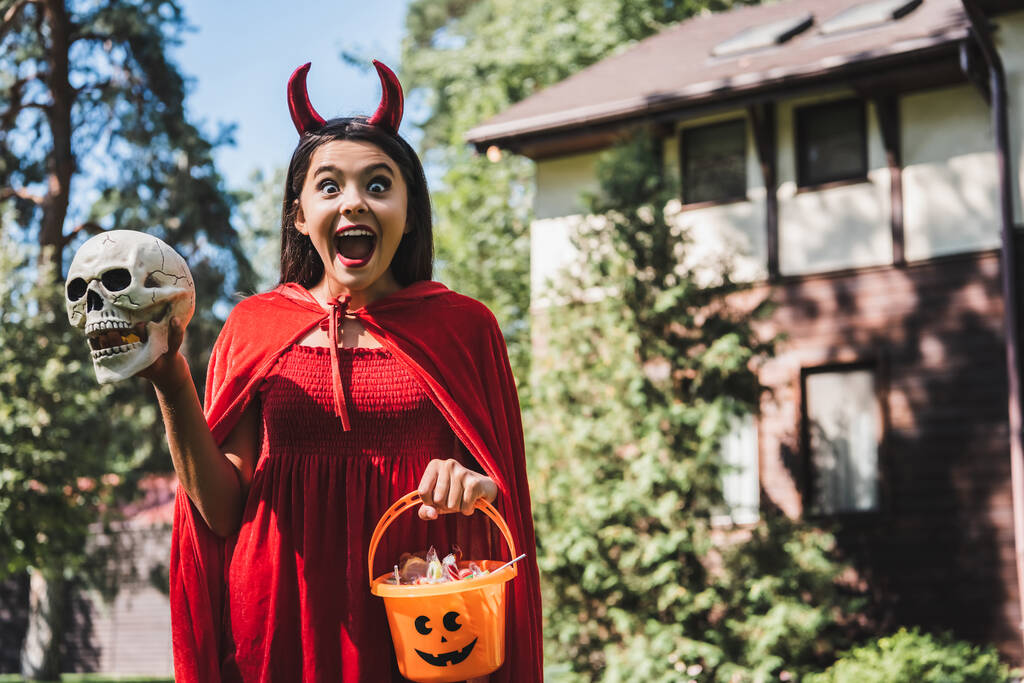 bambino stupito in costume demoniaco urlando mentre teneva teschio e secchio di Halloween vicino al cottage sfocato - Foto, immagini