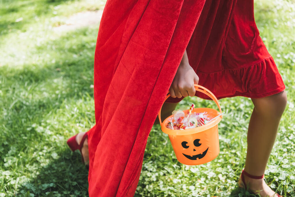 vue recadrée de fille en costume rouge portant seau d'Halloween avec des bonbons tout en marchant sur l'herbe - Photo, image