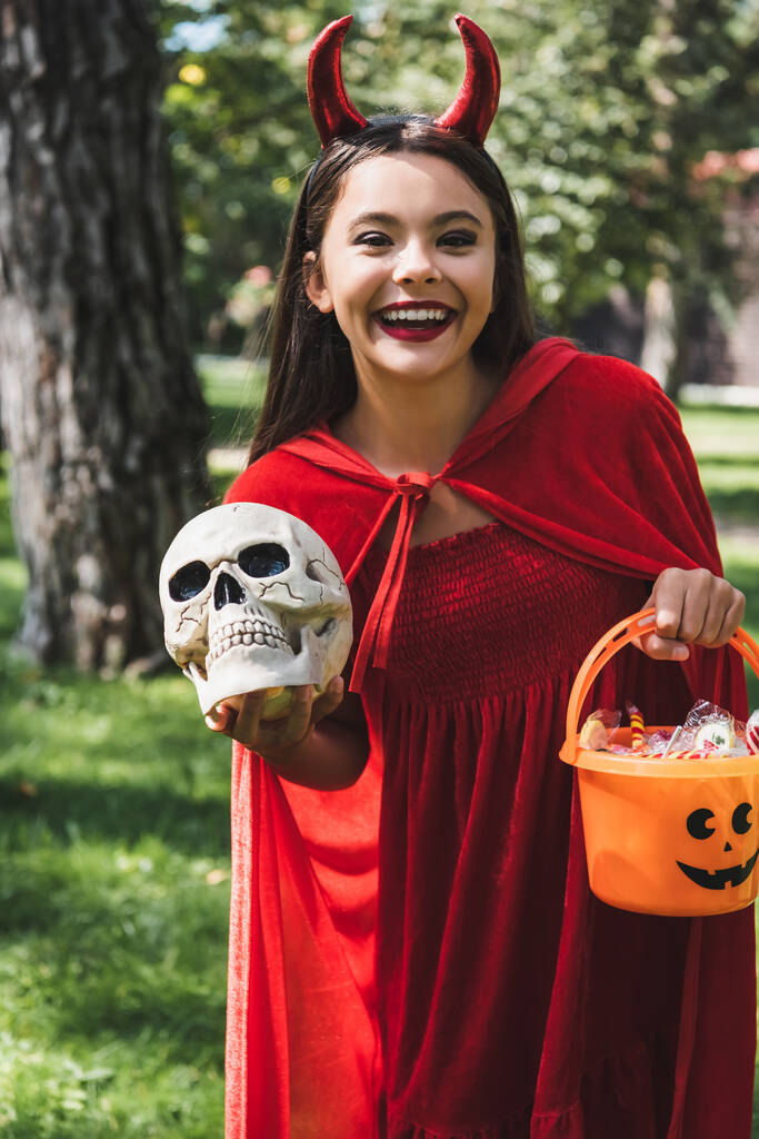 веселая девушка в костюме демона на Хэллоуин держит череп и ведро с конфетками на улице - Фото, изображение