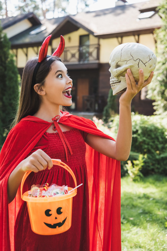 amazed girl in devil halloween costume looking at spooky skull while holding bucket with candies - Photo, Image