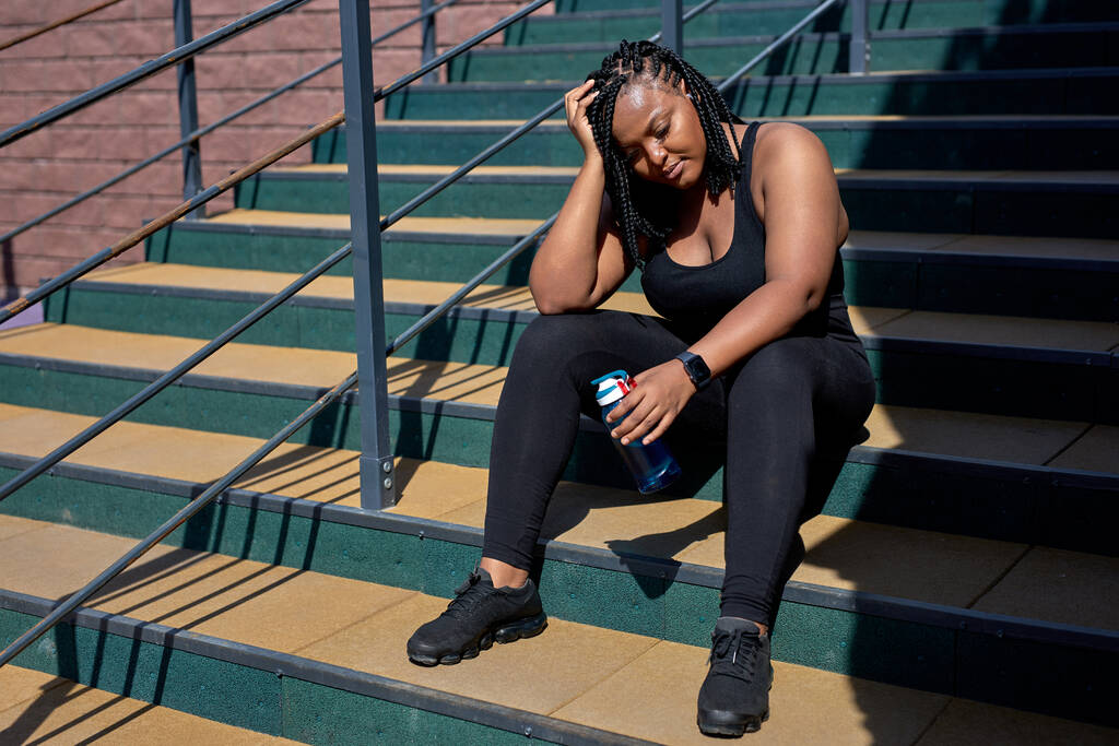 Κουρασμένη Αφρο-Αμερικανίδα με μαύρη φόρμα να ξεκουράζεται ενώ κάθεται στις σκάλες έξω. - Φωτογραφία, εικόνα