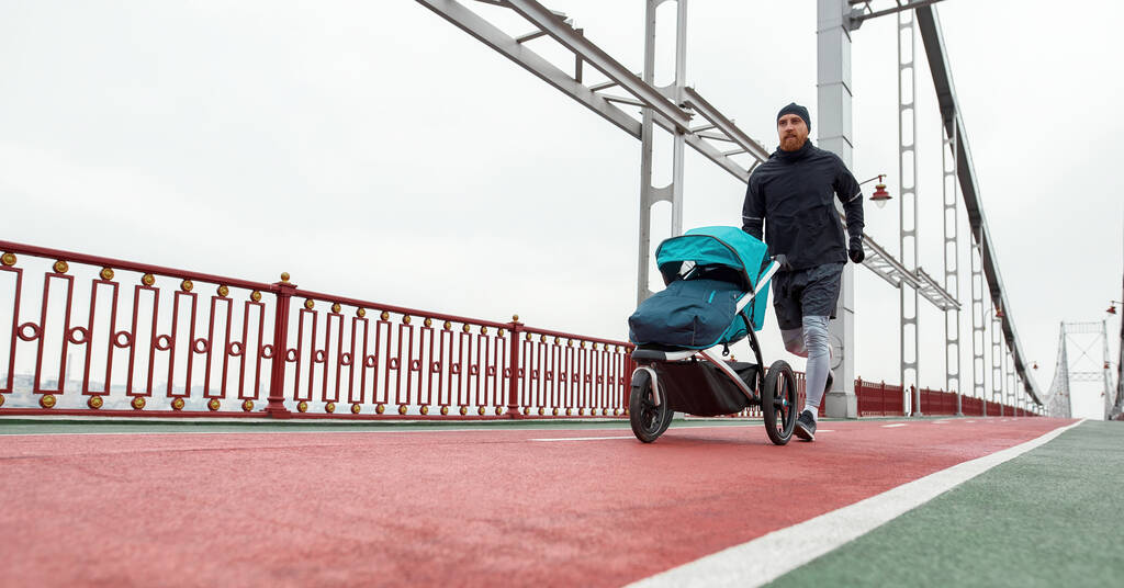 Tiro de comprimento total de pai jovem ativo correndo com uma carruagem de bebê em um dia nublado na cidade - Foto, Imagem