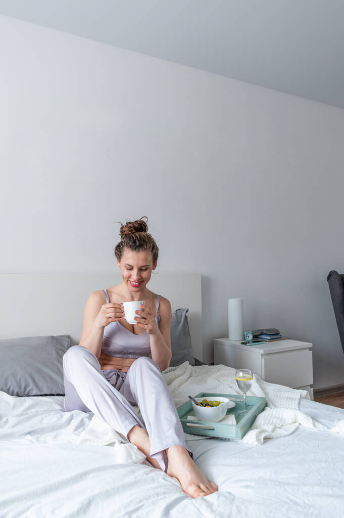 A fiatal nő selyem pizsamában egészséges reggelit reggelizik, miközben az ágyon fekszik a hálószobában. Otthoni jólét koncepció. Egy fiatal nő érzelmi egészsége - Fotó, kép