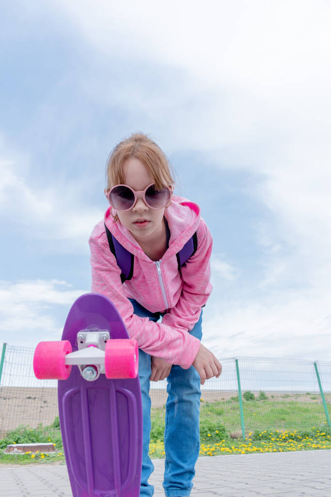 Jovem de jeans e suéter rosa segura um skate na rua. Conceito de estilo de vida adolescente. Espaço para cópia. Mochila roxa nas costas. - Foto, Imagem