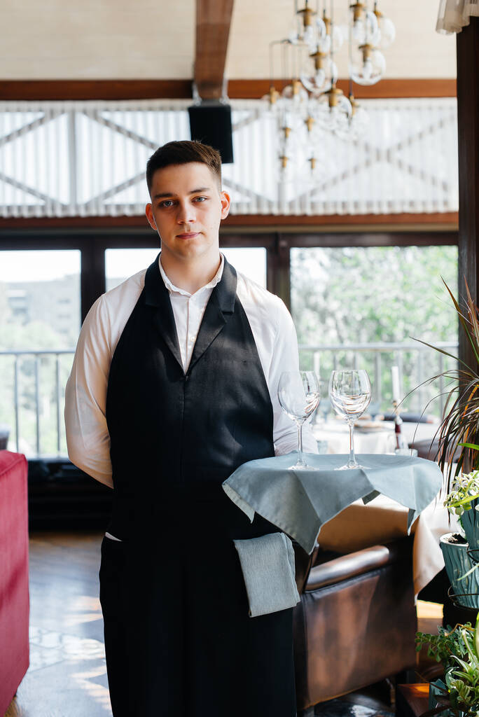 Młody kelner w stylowym mundurze stoi z okularami na tacy w pobliżu stołu w pięknej restauracji z bliska. Aktywność restauracji na najwyższym poziomie. - Zdjęcie, obraz