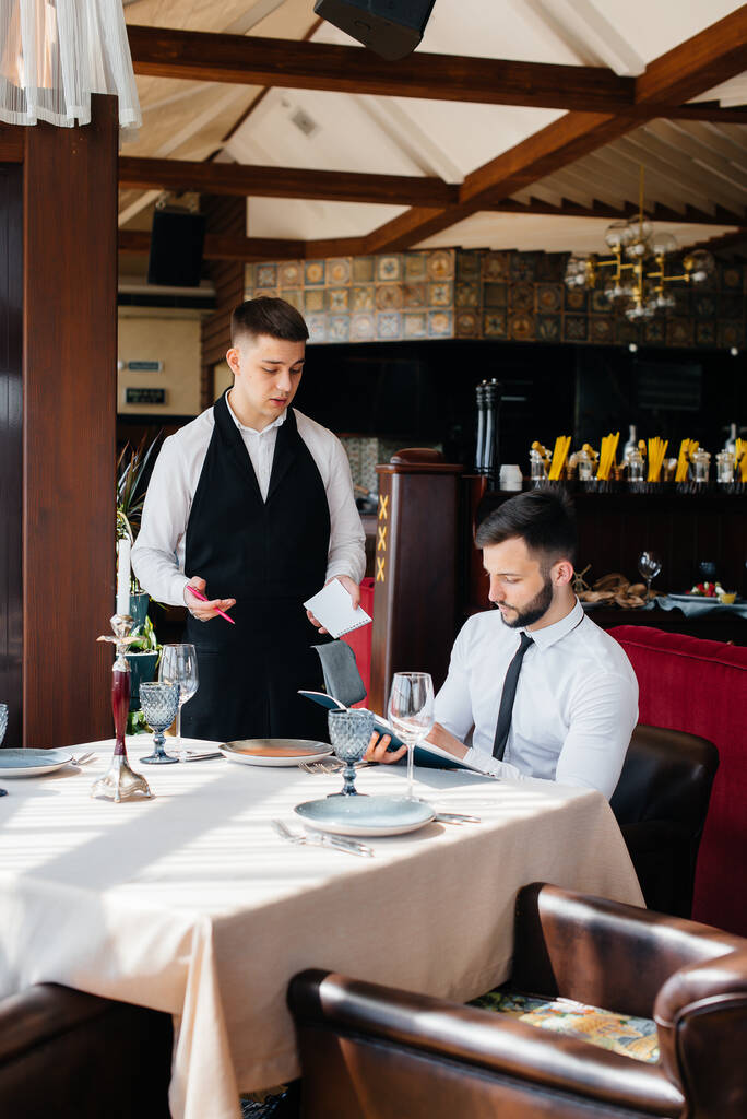 Un joven hombre de negocios en un buen restaurante examina el menú y hace un pedido a un joven camarero en un delantal con estilo. Servicio al cliente. - Foto, Imagen