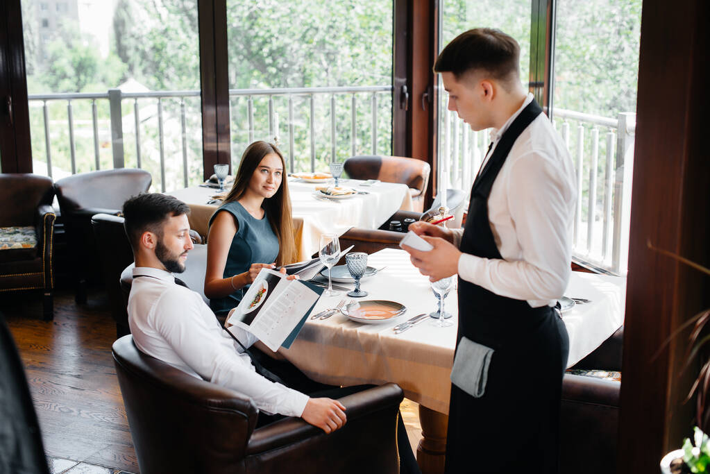 Um garçom elegante serve um jovem casal que veio em uma data em um restaurante gourmet. Atendimento ao cliente no catering. - Foto, Imagem