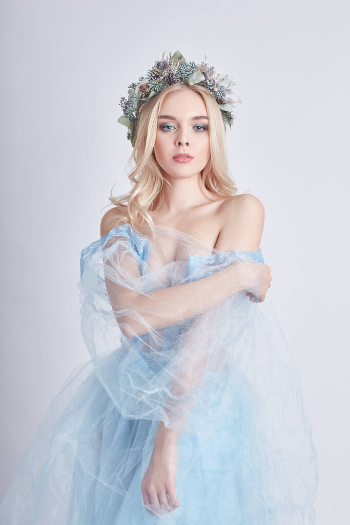 青いエーテルのドレスと白い背景に頭の上に花輪で魅力的な妖精の女性、完璧な肌とメイクを持つ穏やかな神秘的なブロンドの女の子。清潔感、ボディケア、肌 - 写真・画像