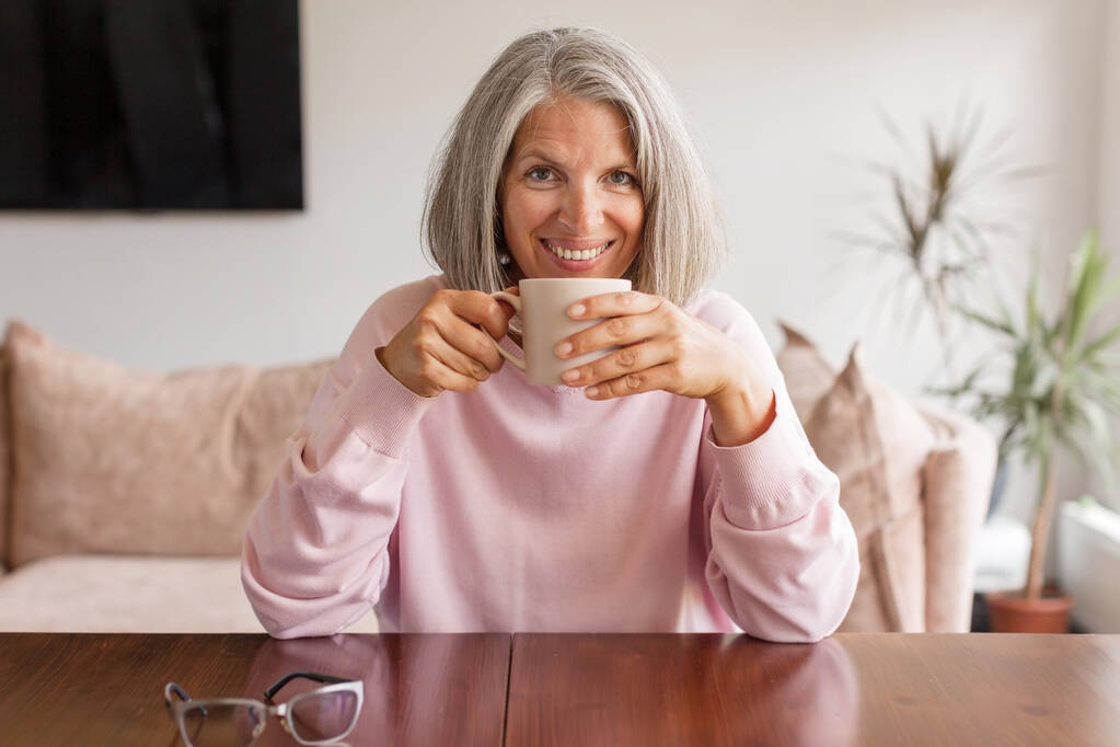 静かで穏やかな中年の白髪の女性が休憩を取り、自宅で熱いお茶のカップで休んでいます。内容魅力的な幸せな成熟した女性はコーヒーと夢を楽しんで - 写真・画像