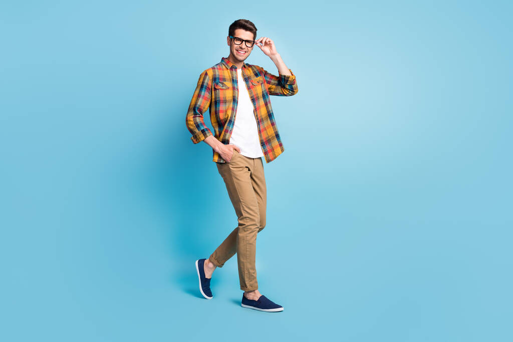 Foto de caballero joven bastante atractivo usar gafas de brazo camisa a cuadros caminando sonriendo aislado fondo de color azul - Foto, imagen