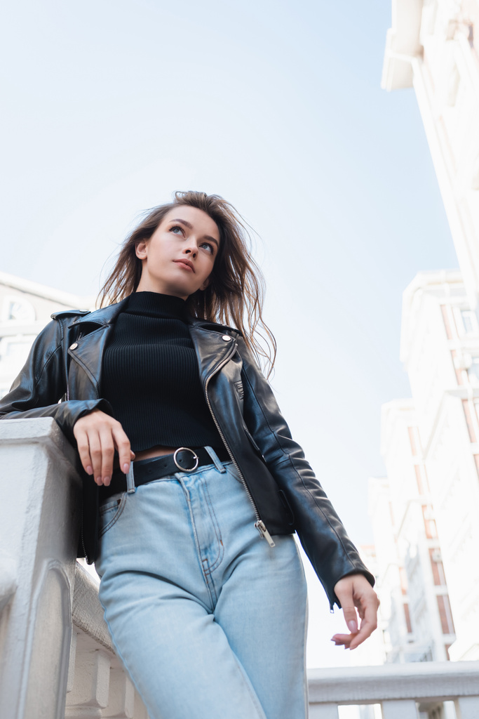 vista de ángulo bajo de la bonita mujer joven en chaqueta de cuero negro y jeans de pie en la calle urbana - Foto, imagen