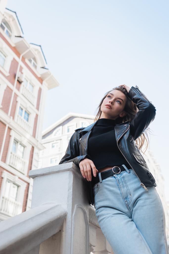 low angle view of young woman in black leather jacket and jeans adjusting hair on urban street - Photo, Image