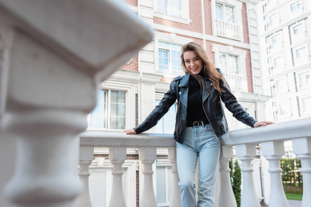 pozitivní mladá žena v černé kožené bundě s úsměvem na městské ulici v Evropě  - Fotografie, Obrázek