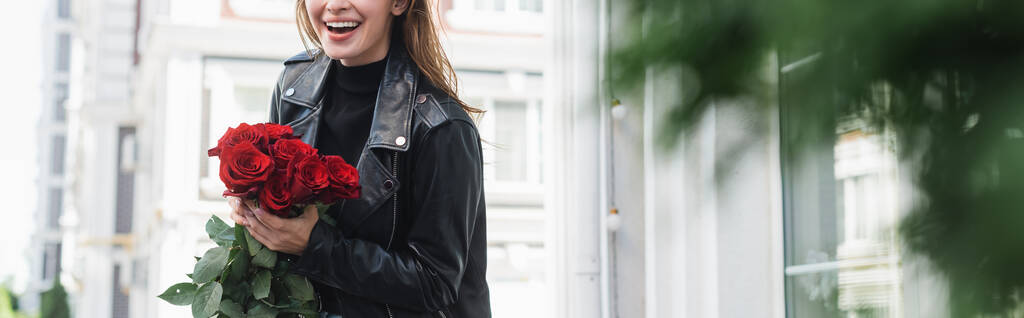 Kırpılmış, deri ceketli, Avrupa 'da şehir sokağında kırmızı güller taşıyan mutlu genç kadın manzarası, afiş. - Fotoğraf, Görsel