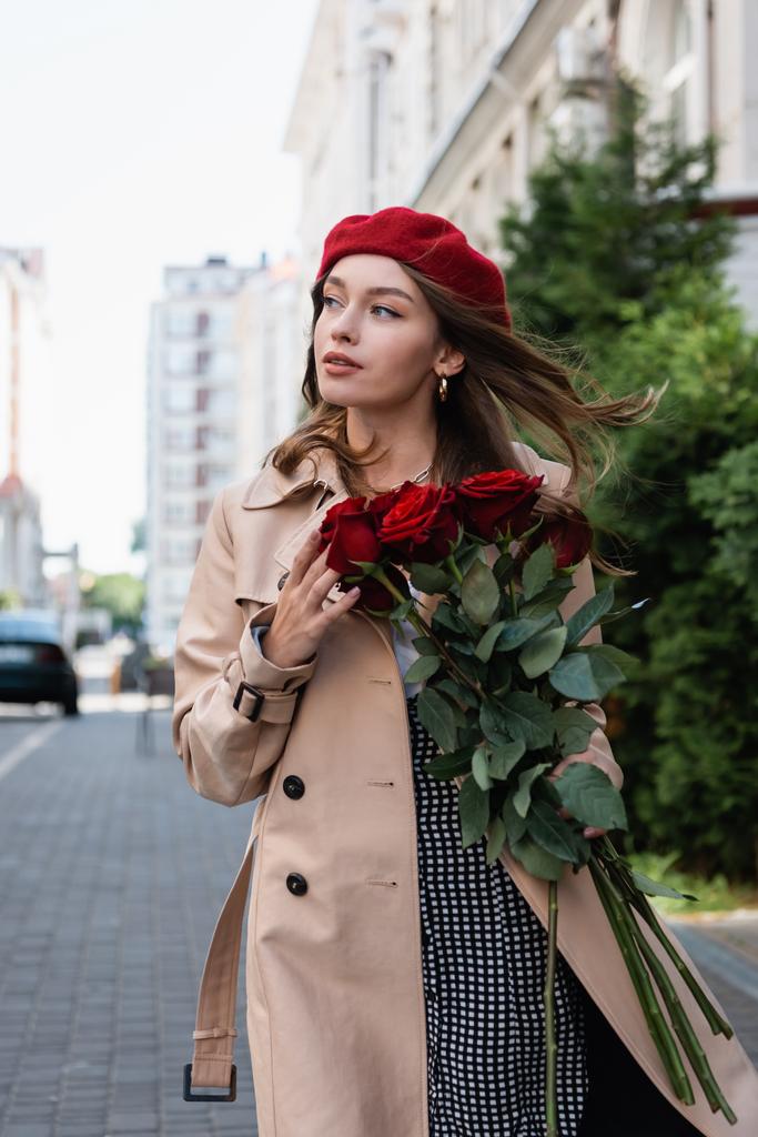 όμορφη γυναίκα σε καμπαρντίνα και κόκκινο μπερέ κρατώντας μπουκέτο τριαντάφυλλα στο αστικό δρόμο της Ευρώπης  - Φωτογραφία, εικόνα