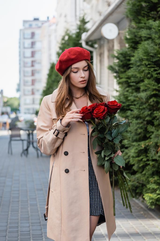 csinos nő ballonkabátban és piros sapkában nézi csokor rózsa a városi utcán Európa  - Fotó, kép