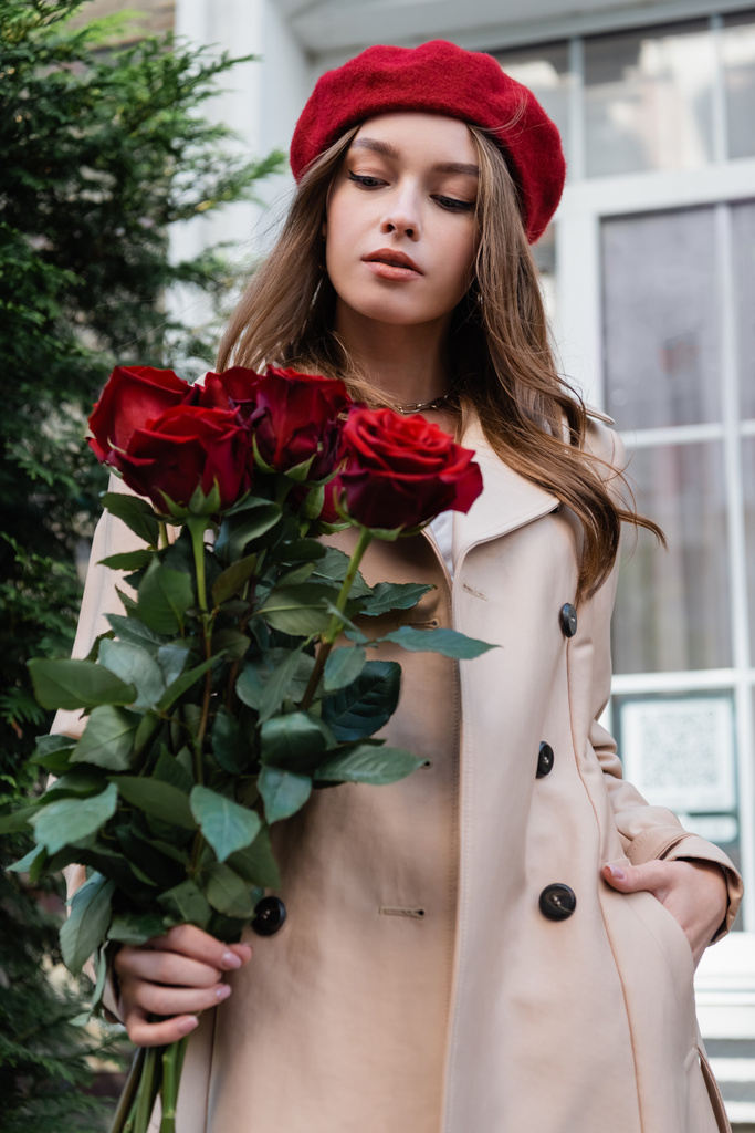 bella donna in trench e berretto rosso guardando mazzo di rose vicino edificio  - Foto, immagini