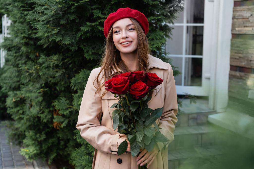 χαρούμενη γυναίκα με καμπαρντίνα και κόκκινο μπερέ κρατώντας τριαντάφυλλα κοντά στο κτίριο  - Φωτογραφία, εικόνα