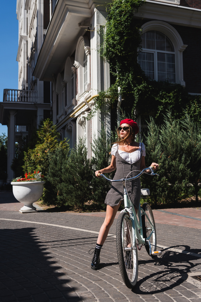 longitud completa de bonitas gafas de sol de mujer, boina roja y vestido de pie cerca de la bicicleta en la calle urbana en Europa  - Foto, Imagen