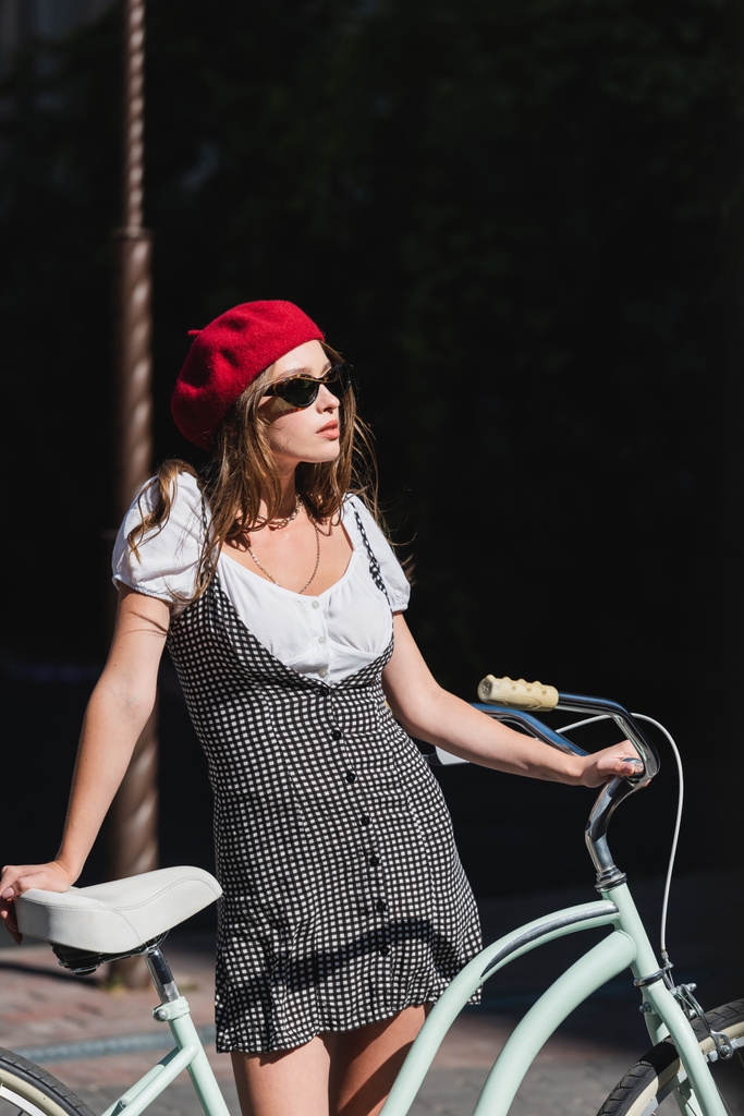 młoda kobieta w okularach przeciwsłonecznych, czerwonym berecie i sukience stojącej na rowerze  - Zdjęcie, obraz