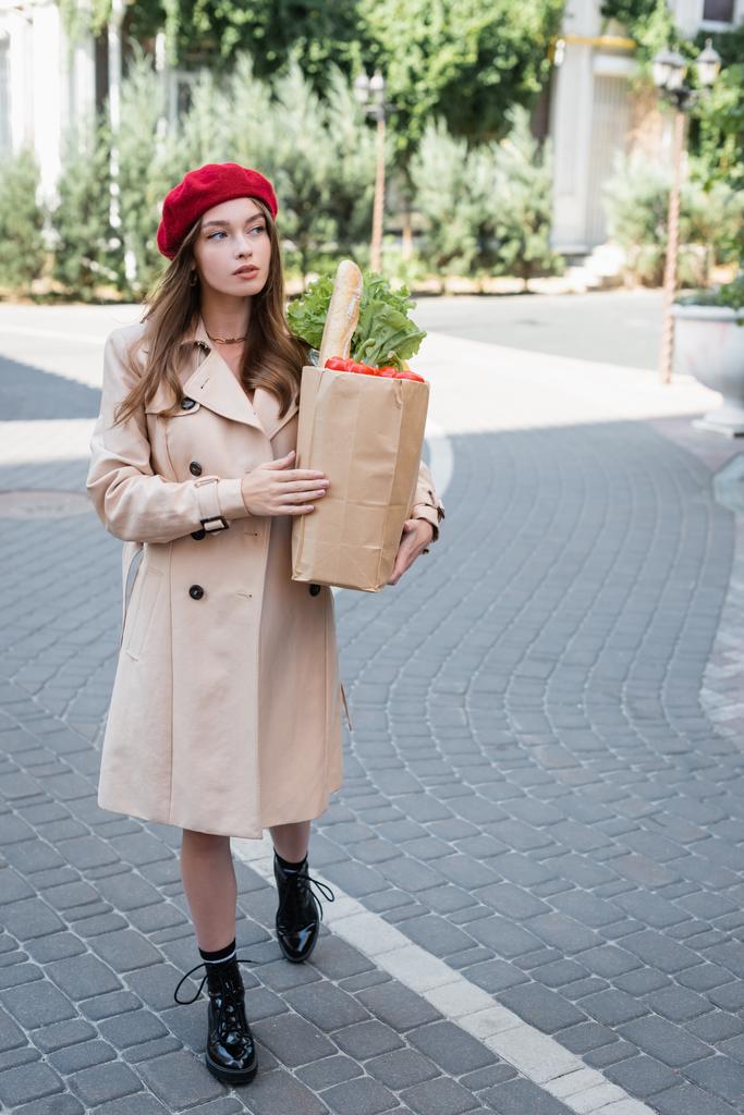 volledige lengte van jonge vrouw in beige trench jas en rode baret holding papieren zak met boodschappen op stedelijke straat  - Foto, afbeelding