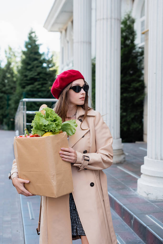 mladá žena v plášti, slunečních brýlích a červeném baretu drží papírový sáček s potravinami v blízkosti budovy  - Fotografie, Obrázek