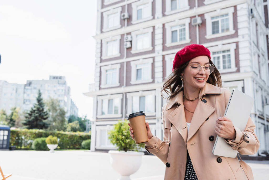 positive freelancer in red beret and beige trench coat holding laptop and paper cup of street  - Photo, Image
