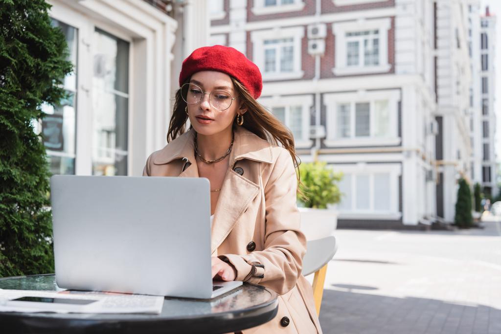 muito freelancer em óculos, boina vermelha e casaco bege usando laptop no terraço do café  - Foto, Imagem