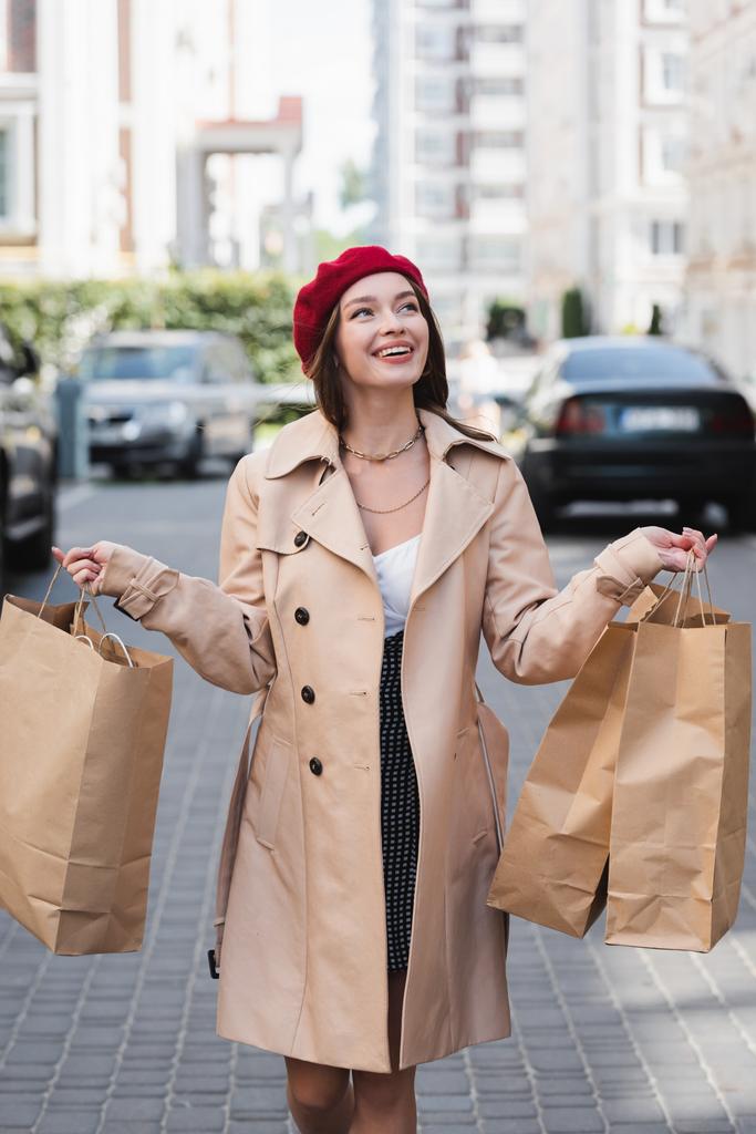 mujer alegre en boina roja y gabardina beige sosteniendo bolsas de compras en la calle urbana  - Foto, Imagen
