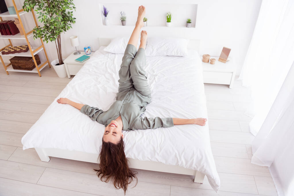 Fénykép édes vicces fiatal nő szürke hálóing fekvő ágy mosolygó emelkedő lábak belsejében beltéri otthoni szobában - Fotó, kép