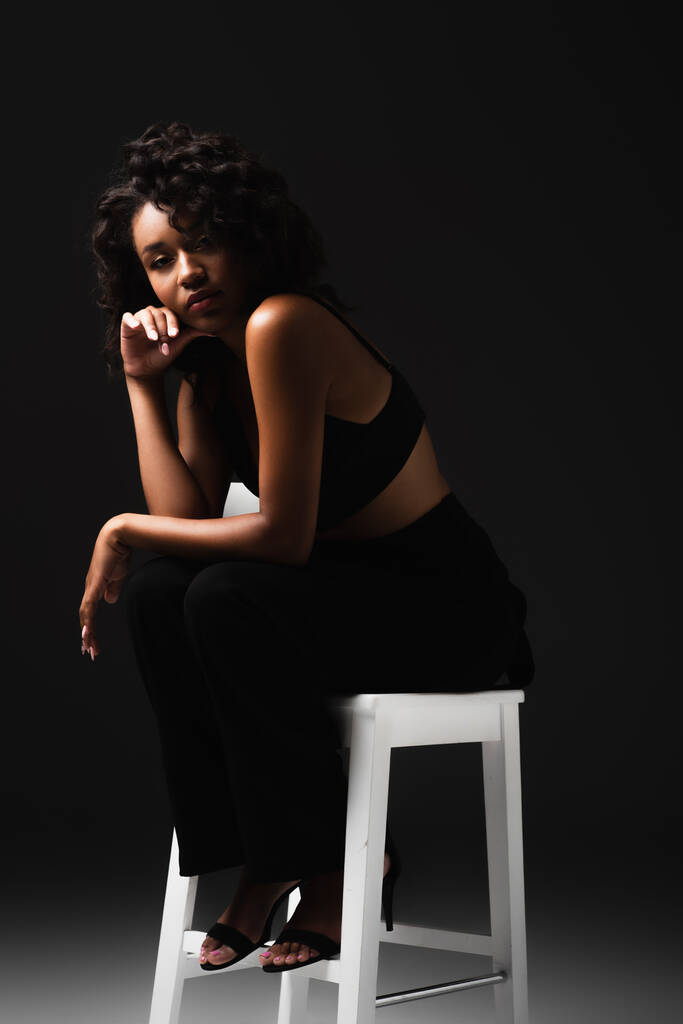 σέξι αφροαμερικανίδα γυναίκα σε σατέν κορυφή καλλιέργεια κάθεται σε λευκή καρέκλα σε μαύρο  - Φωτογραφία, εικόνα