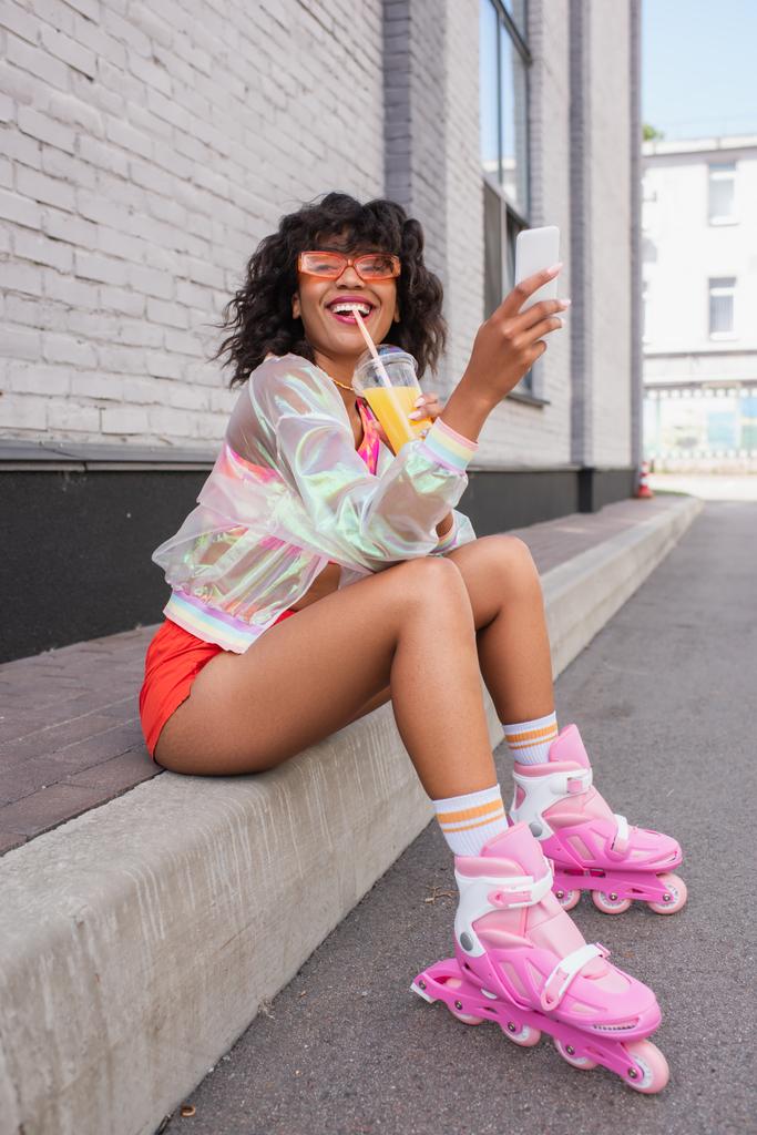 volledige lengte van glimlachende Afro-Amerikaanse vrouw in zonnebril en rolschaatsen drinken van sinaasappelsap terwijl het houden van mobiele telefoon - Foto, afbeelding