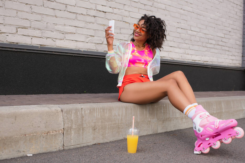 full length of happy african american woman in sunglasses and roller skates using smartphone on urban street - Photo, Image