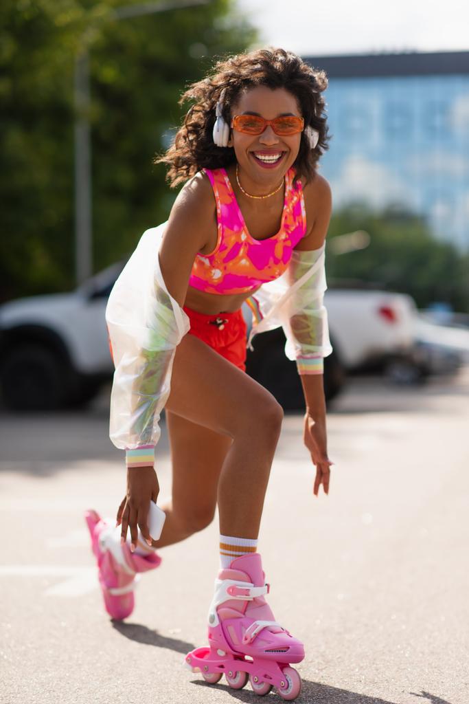 uśmiechnięta Afroamerykanka w słuchawkach bezprzewodowych jeżdżąca na wrotkach trzymając komórkę na zewnątrz  - Zdjęcie, obraz